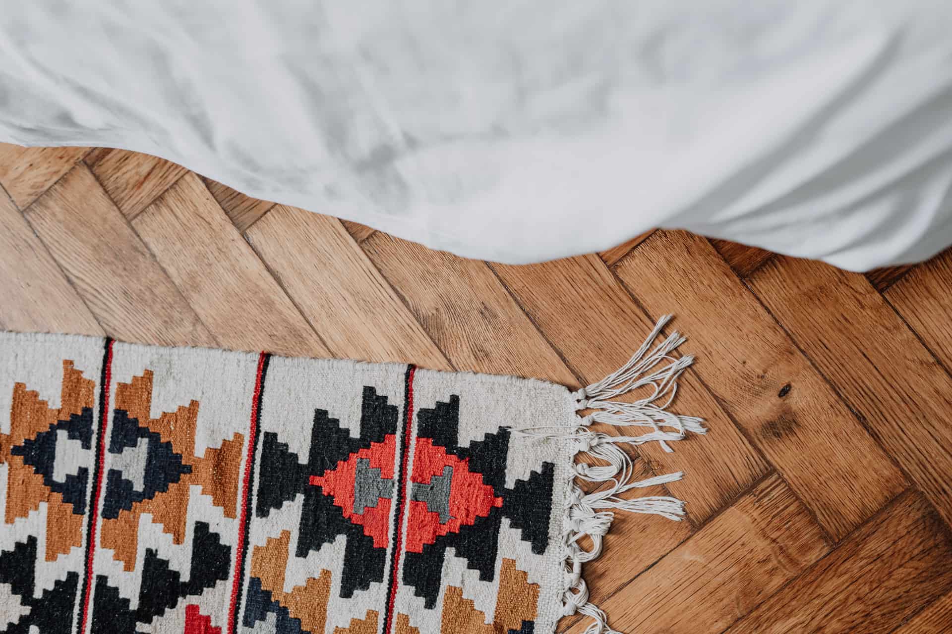 A small flatweave accent rug with tasseled ends on a wood floor beside a bed